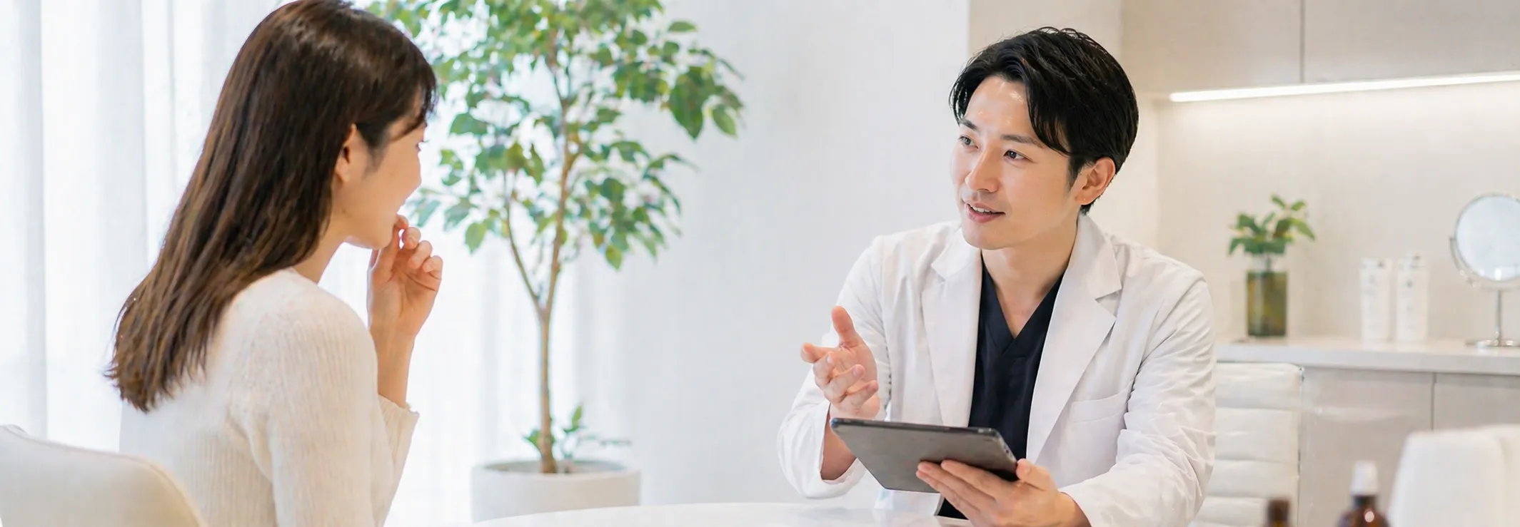 医師がタブレットを持ちながら患者に説明している診察シーン。明るいオフィス環境に観葉植物が配置されている